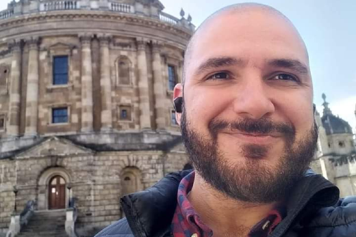 Imagem: Homem careca com barba sorrindo para a câmera em um local histórico. Ele está usando uma jaqueta preta e fones de ouvido. Ao fundo, um prédio de arquitetura clássica com fachada de pedra e grandes janelas, sugerindo um ambiente de importância histórica ou acadêmica