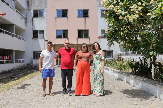 Imagem: quatro pessoas de mãos dadas posam em pé para uma fotografia, em frente a um prédio 