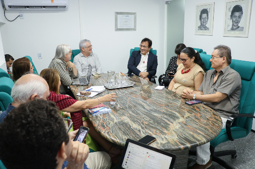 Nesta imagem, um grupo de pessoas está sentado ao redor de uma mesa de reunião oval de mármore, em um ambiente de escritório. Alguns dos participantes parecem discutir assuntos sérios, enquanto outros observam. Há copos e uma bandeja com água sobre a mesa. As cadeiras são acolchoadas e de cor verde. Na parede ao fundo, há retratos emoldurados, além de um aparelho de ar-condicionado e uma placa emoldurada no centro da parede.