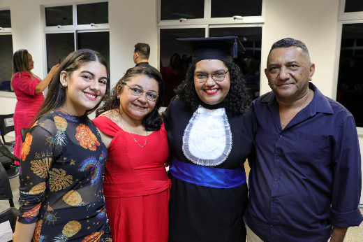 Imagem: quatro pessoas posam sorridentes para a foto em um ambiente fechado. Ao centro, uma mulher usa beca e capelo de formatura, simbolizando sua conquista. Ela está ladeada por duas mulheres, uma de vestido estampado e outra de vestido vermelho, e por um homem de camisa azul. Todos parecem felizes, celebrando juntos um momento especial.