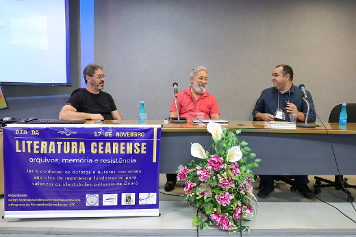 Imagem: a imagem mostra uma mesa redonda em um evento sobre literatura cearense, com três homens sentados ao redor de uma mesa em um auditório.  À esquerda, um homem de óculos, vestindo uma camiseta preta e calça escura, está sentado e sorrindo levemente. Ao centro, um homem de cabelos grisalhos e barba branca curta, vestindo uma camisa social rosa e calça preta, está sentado com as mãos apoiadas na mesa. Ele também sorri, transmitindo simpatia. À direita, um terceiro homem, de cabelo curto, usando uma camisa de manga longa azul escura e um crachá pendurado no pescoço, conversa com os outros enquanto segura um livro. Na mesa, há garrafas de água e papéis. À frente, um banner roxo com letras amarelas e brancas traz o texto sobre o evento