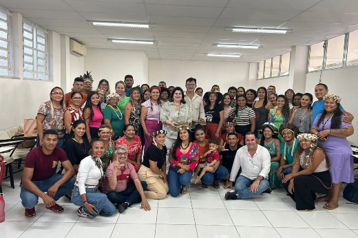 Imagem: A imagem mostra um grande grupo de pessoas reunidas em uma sala ampla e iluminada, com paredes brancas e janelas altas. A maioria das pessoas está de pé, enquanto algumas estão agachadas na frente. Algumas mulheres estão usando cocares e adereços indígenas. No centro, uma mulher e um homem seguram um objeto, possivelmente um troféu ou homenagem. Todos aparentam estar felizes e participando de um evento ou celebração. O piso é de cerâmica branca e o teto possui iluminação com lâmpadas fluorescentes.