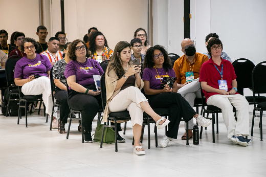 Imagem: A imagem mostra um grupo de pessoas sentadas em cadeiras, assistindo a um evento em um espaço interno iluminado. A maioria dos participantes está disposta em fileiras e olha para frente, sugerindo que estão atentos a uma apresentação ou painel.  Na frente, destacam-se algumas pessoas: Duas mulheres usando camisetas roxas com o texto "CULTURA UFC" e calças claras. Uma mulher ao centro veste roupas claras e está mexendo em um celular. Outras duas pessoas usam trajes casuais; uma delas tem um crachá azul. Mais ao fundo, é possível ver outras pessoas, algumas de pé, e um homem de máscara preta. A atmosfera do evento parece formal, mas descontraída, sugerindo uma atividade relacionada a temas acadêmicos ou culturais promovidos pela Universidade Federal do Ceará (UFC)