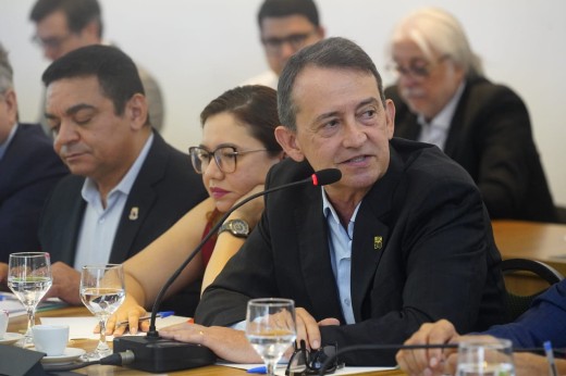 A imagem mostra uma mesa de reunião com várias pessoas sentadas lado a lado, em ambiente interno e formal. O foco está em um homem de terno preto e camisa azul clara, falando ao microfone. À sua direita, uma mulher com óculos e expressão atenta observa a reunião. Também há outros participantes ao fundo, com copos e garrafas de água sobre a mesa. O ambiente é iluminado e sugere um encontro profissional ou institucional.