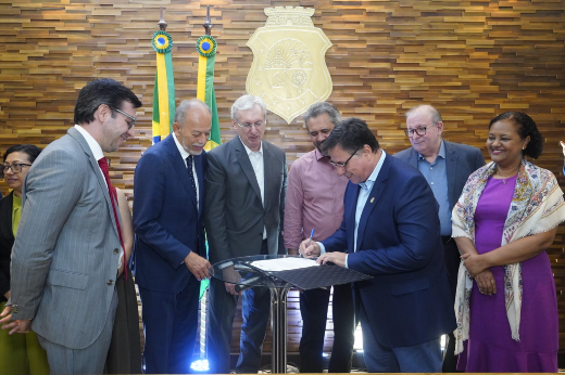 A imagem mostra um grupo de sete pessoas reunidas em um evento formal. Eles estão em frente a uma parede de madeira com o brasão do estado do Ceará no centro, entre duas bandeiras do Brasil. Uma pessoa está assinando um documento em um púlpito transparente, enquanto as outras observam e sorriem. Todos estão vestidos formalmente, com roupas sociais e ternos. O ambiente está bem iluminado, indicando ser um local de cerimônia oficial.