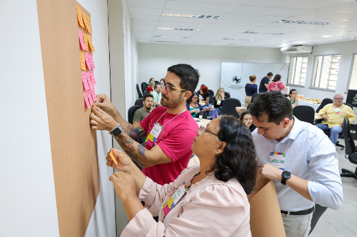 Imagem: A imagem mostra um grupo de pessoas participando de uma atividade colaborativa em um ambiente interno, provavelmente uma sala de reunião ou treinamento. No centro da cena, três pessoas estão interagindo com um painel coberto de papel kraft, fixando post-its coloridos (rosas, laranjas e amarelos). Um dos participantes, à esquerda, veste uma camiseta rosa e óculos, enquanto outro, à direita, veste uma camisa branca e segura o papel para facilitar a fixação dos post-its. A terceira pessoa, no centro, também contribui com os post-its.  Ao fundo, outras pessoas estão sentadas ao redor de mesas, aparentemente participando da mesma dinâmica. O ambiente é bem iluminado, com janelas grandes à direita e banners institucionais visíveis ao fundo, incluindo um com o logo da Universidade Federal do Ceará (UFC). Sobre uma mesa ao fundo, há o que parece ser um coffee break com lanches e bebidas. O cenário transmite uma atmosfera de trabalho colaborativo e interação.