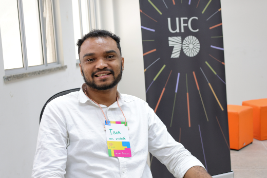 Imagem: A imagem mostra um homem sentado, olhando para a câmera e sorrindo. Ele veste uma camisa branca e usa um crachá colorido pendurado no pescoço, onde está escrito "Igor" e "UFC Itapajé". Ao fundo, há um banner preto com o logo "UFC 70", decorado com linhas coloridas irradiando do centro, e blocos de assento laranja no canto direito. À esquerda, vê-se parte de uma janela com luz natural entrando no ambiente. A expressão do homem transmite simpatia, e o contexto parece ser um evento institucional ou acadêmico.