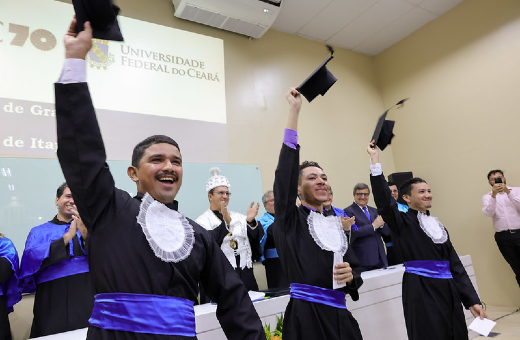 Imagem: três estudantes vestindo beca levantam capelos de cor preta e comemoram a formatura