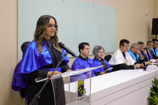 Imagem: moça de óculos e cabelos castanhos vestindo beca preta e túnica azul discursa em púlpito