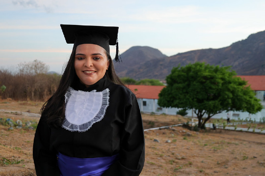 Imagem: uma mulher em ambiente externo veste trajes de formatura. Ela usa uma beca preta com detalhes brancos no peito e uma faixa azul na cintura, além de um capelo preto com uma franja pendente ao lado. Ela sorri levemente, com os cabelos soltos, e está em frente a uma paisagem composta por um terreno seco, uma árvore verde e, ao fundo, um prédio branco com telhado vermelho e montanhas sob um céu azul claro.