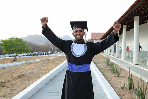 Imagem: um homem negro em um ambiente externo celebra sua formatura. Ele veste uma beca preta com detalhes em branco no peito e uma faixa azul na cintura, além de um capelo preto. Ele sorri, com os braços erguidos em sinal de vitória. Ao fundo, há um prédio com colunas brancas, uma vegetação árida e montanhas ao longe, sob um céu claro.