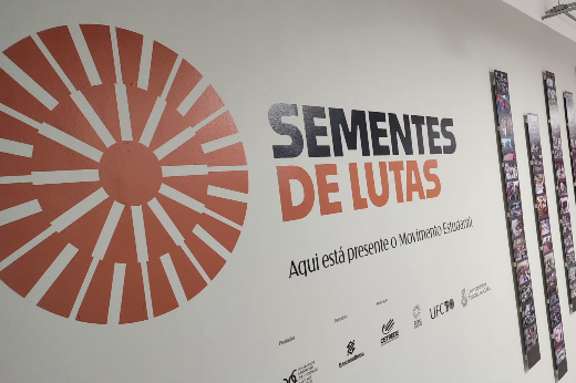 Imagem: uma parede com a inscrição "Sementes de Lutas" em letras grandes, onde "Sementes" está em preto e "de Lutas" em laranja. Abaixo, lê-se: "Aqui está presente o Movimento Estudantil". Ao lado esquerdo, há um símbolo em forma de círculo laranja com raios que se expandem para fora, remetendo a um gráfico radial ou ao conceito de união e difusão de ideias.  No lado direito da imagem, colunas verticais exibem fotografias históricas de lutas e manifestações relacionadas ao movimento estudantil, organizadas em sequência.