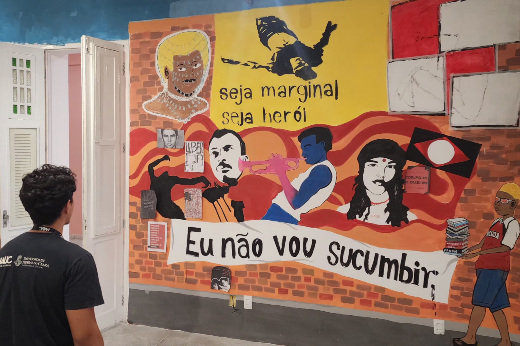 Imagem: um mural artístico em uma parede com diversos elementos visuais e mensagens. O fundo é composto por tijolos pintados, e sobre eles há imagens e textos em diferentes estilos artísticos. No centro, há a frase "Eu não vou sucumbir" escrita em preto sobre um fundo branco. Acima, lê-se "Seja marginal, seja herói" em letras pretas sobre um fundo amarelo.  O mural inclui representações de pessoas, como um homem tocando trompete, rostos estilizados, e um jovem negro à direita carregando uma pilha de livros. Há também referências a símbolos culturais e políticos, incluindo cartazes e bandeiras. No canto esquerdo inferior, um homem de costas observa a arte.