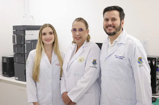 Imagem: A imagem mostra três pessoas vestindo jalecos brancos em um ambiente que parece ser um laboratório científico. À esquerda, uma mulher de cabelos loiros longos sorri. No centro, uma mulher de óculos, cabelos presos e um sorriso discreto está entre os outros dois. À direita, um homem de barba e cabelo curto sorri para a câmera. No fundo, há equipamentos laboratoriais e bancadas, indicando que eles são pesquisadores ou profissionais da área de saúde ou ciência