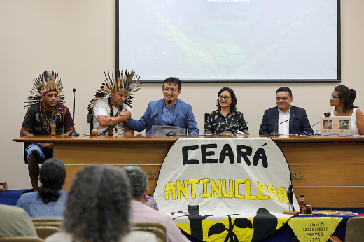 Imagem: A imagem mostra um evento com várias pessoas sentadas em uma mesa, incluindo representantes indígenas com cocares, acompanhados de outros participantes. À frente da mesa, há uma faixa com os dizeres "Ceará Antinuclear". Parece ser uma reunião ou debate relacionado a temas ambientais e energéticos, com foco na questão nuclear.