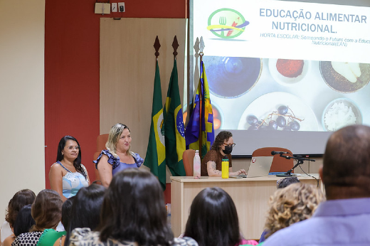 Imagem: Na imagem, há um ambiente de apresentação em um auditório. Três mulheres estão em frente à plateia, próximas a uma mesa e a um telão. No telão, lê-se: "Educação Alimentar e Nutricional" e "Horta Escolar: Semeando o Futuro com a Educação Alimentar e Nutricional (CECANE/UFV)". Ao fundo, há bandeiras do Brasil, de um estado e de um município. A plateia é composta por várias pessoas, sentadas e atentas à apresentação.