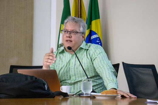 Imagem: um homem de cabelo grisalho, óculos e camisa listrada verde, sentado à mesa durante uma fala. Ele gesticula com a mão direita e está diante de um notebook aberto, com um microfone posicionado à sua frente. Sobre a mesa, há um copo com água, uma xícara, e um prato com pedaço de bolo. Ao fundo, estão visíveis bandeiras, incluindo a do Brasil.