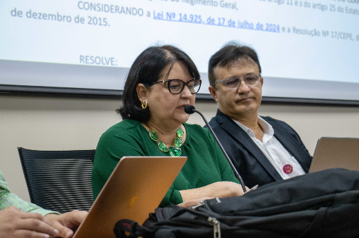 Imagem: mulher de cabelos pretos e curtos, com óculos, blusa verde e colar de pedras grandes verdes aparece falando ao microfone, sentada atrás de uma mesa; ao seu lado, aparece um homem de cabelos curtos e lisos, óculos , camisa social branca e jaqueta do paletó, olhando para um papel que a mulher segura nas mãos