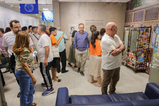 Imagem: um grupo de pessoas está reunido em um espaço que parece ser uma exposição ou museu. Algumas pessoas estão paradas observando detalhes nas paredes, que incluem textos e fotografias. No lado direito, há uma máquina ou armário decorado com adesivos coloridos. Um sofá azul está em primeiro plano. O ambiente é bem iluminado e conta com painéis no teto e paredes claras, criando um clima de interação e interesse.