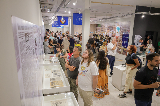 Imagem:  espaço de exposição interna, com várias pessoas caminhando e observando objetos expostos em vitrines de vidro. Ao fundo, há bandeiras azuis com o logotipo da UNE (União Nacional dos Estudantes). O ambiente parece ser iluminado com luz branca, e as paredes estão decoradas com painéis exibindo imagens e textos históricos. O público é diverso, com pessoas de várias idades, algumas interagindo entre si, enquanto outras estão concentradas nos itens expostos. O local transmite uma atmosfera de interesse cultural e histórico.