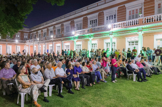 Imagem:  evento ao ar livre durante a noite, realizado em frente a um grande prédio histórico de fachada rosa e branca com arquitetura colonial, iluminado por refletores verdes. Um grande público está sentado em cadeiras brancas de plástico, organizado em fileiras sobre o gramado. Entre os participantes, há pessoas de diferentes idades, com algumas em pé e outras conversando. O ambiente é iluminado e festivo, transmitindo a sensação de uma celebração ou encontro cultural.