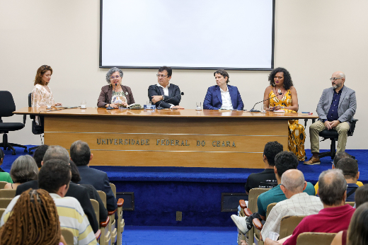 Imagem: um grupo de seis pessoas, entre homens e mulheres, aparece sentado atrás de uma mesa comprida de madeira, no palco de um auditório. Uma das mulheres fala ao microfone. Atrás deles aparece uma tela grande de projeção. Na frente da mesa lê-se o nome Universidade Federal do Ceará. Á frente do grupo estão pessoas da plateia