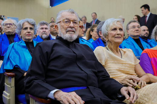 Imagem: Um grupo de pessoas sentadas, aparentando participar de uma cerimônia formal. No primeiro plano, um homem idoso de barba branca, vestindo uma roupa preta, e uma mulher idosa ao seu lado, com cabelo branco curto e um vestido claro. Atrás deles, várias outras pessoas, algumas usando capas ou mantos azuis, também estão sentadas. O ambiente é iluminado e parece ser um auditório ou sala de conferências, com outras pessoas em pé ao fundo