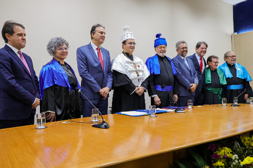 Imagem: um grupo de nove pessoas posa para foto, de pé, atrás de uma mesa comprida de madeira, dentro de um auditório. Algumas trajam vestes talares, nas cores azul e verde, outras vestem terno e gravata