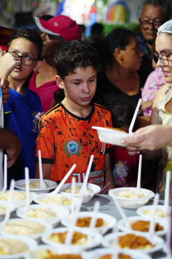 Imagem: grupo de pessoas em um evento de distribuição de alimentos. No centro, há uma criança usando uma camiseta laranja, olhando para uma mesa cheia de pratos descartáveis com comida e colheres. Ao lado dele, outros jovens e adultos estão em fila, esperando para serem servidos. Uma mulher está entregando um prato a alguém. O ambiente parece movimentado e acolhedor, com diversas pessoas participando da atividade em segundo plano.