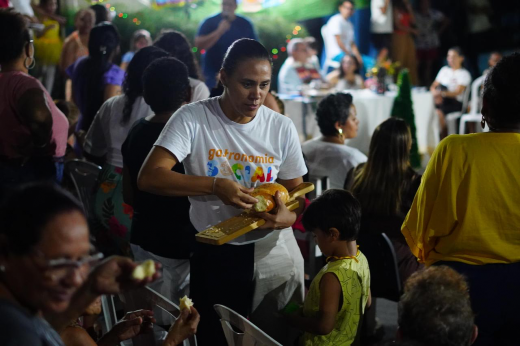 Imagem: momento em um evento ao ar livre, provavelmente uma ação comunitária. No centro, uma mulher com uma camiseta escrita "Gastronomia Social" serve pão para pessoas ao seu redor. Ela segura uma tábua de madeira com os pães enquanto distribui porções. O ambiente está cheio de pessoas sentadas e em pé, e ao fundo há mesas decoradas e luzes, criando uma atmosfera festiva e acolhedora. Crianças e adultos estão presentes no local.