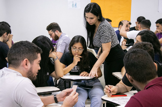 Imagem: A imagem mostra uma sala de aula com várias pessoas sentadas em cadeiras, organizadas em pequenos grupos. Elas parecem estar participando de uma atividade colaborativa. No centro da cena, uma mulher de cabelos longos e pretos, vestida com uma blusa estampada e calça preta, está em pé, inclinando-se para ajudar uma estudante. A estudante está concentrada, segurando um lápis e um caderno. Ao redor, outros participantes também discutem e escrevem, demonstrando envolvimento na atividade. O ambiente é iluminado e descontraído, sugerindo um clima de aprendizado e interação.