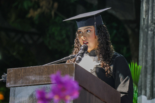 Imagem: uma jovem mulher de cabelos castanhos e cacheados aparece vestindo chapéu de formatura e beca, atrás de um púlpito, falando ao microfone