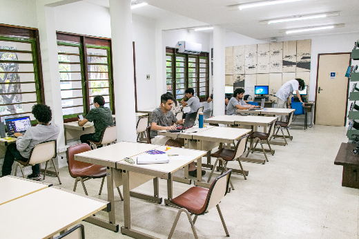 Imagem: A imagem mostra uma sala de estudos com várias mesas e cadeiras distribuídas uniformemente. Algumas pessoas estão sentadas, utilizando computadores ou lendo. A sala tem janelas com persianas horizontais de madeira, permitindo a entrada de luz natural. Ao fundo, há uma pessoa de pé organizando algo próximo a uma prateleira ou mesa com computadores. A iluminação é complementada por lâmpadas fluorescentes no teto. O ambiente parece ser uma biblioteca ou espaço de estudo coletivo.