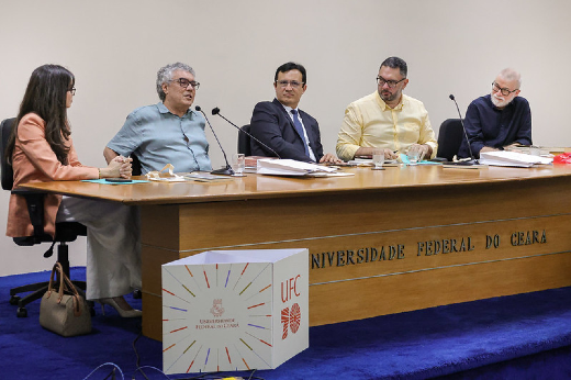 Imagem: Cinco pessoas estão sentadas atrás de uma mesa de madeira em um auditório da Universidade Federal do Ceará (UFC). A mesa tem o nome da instituição gravado na frente. À esquerda, uma mulher de óculos, vestindo um blazer salmão, é a vice-diretora da Editora UFC, Juliana Diniz. Ela escuta um homem de cabelos grisalhos e óculos, que está falando e gesticulando com a mão direita, o escritor Flávio Paiva. Ao lado dele, um homem de terno e gravata olha atentamente para ele. É o reitor da UFC, Custódio Almeida. O quarto participante, o diretor da Editora UFC, Cavalcante Júnior, veste uma camisa amarela de mangas compridas e também observa a conversa. À direita, um homem de barba branca e óculos, vestindo uma camisa preta, inclina-se levemente e parece sorrir. É o escritor Lira Neto. Na frente da mesa, há um painel comemorativo dos 70 anos da UFC. O ambiente tem um fundo claro e neutro.