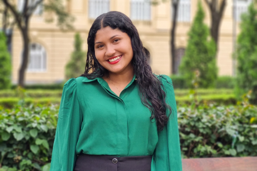 Imagem: A imagem mostra uma jovem sorridente com cabelos longos e ondulados, vestindo uma camisa verde de mangas compridas com detalhes levemente bufantes. Ela também usa uma saia ou calça preta. Ao fundo, há um jardim bem cuidado com arbustos, árvores e grama, além de um edifício de cor clara com janelas grandes e detalhes arquitetônicos clássicos. O cenário é ao ar livre, em um ambiente iluminado e agradável.