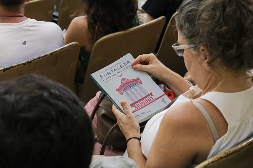 Imagem: Uma mulher, sentada na plateia de um auditório, segura em suas mãos um livro, enquanto olha para a capa. Na capa aparece o título Fortaleza Poéticas do Passado, com a imagem da fachada de uma casa antiga, nas cores vermelho e cinza claro