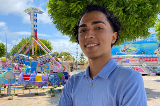 Imagem: A imagem mostra um jovem com cabelo cacheado curto e sobrancelhas bem definidas, sorrindo levemente. Ele veste uma camisa azul clara e está em um ambiente externo durante o dia. Ao fundo, há um parque de diversões com brinquedos coloridos, incluindo um carrossel e estruturas decoradas com imagens alegres. Árvores verdes também estão presentes, oferecendo sombra. O clima parece ensolarado, com céu azul e poucas nuvens.