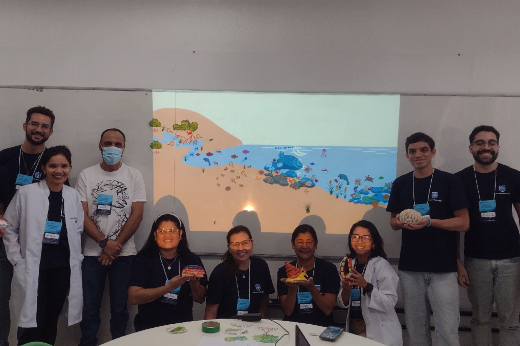 Imagem: Um grupo de nove pessoas em uma sala de aula, posando para a foto. Ao fundo, um projetor exibe uma ilustração de um ambiente marinho com praia, corais e diversos animais aquáticos. Algumas pessoas estão em pé, enquanto outras estão sentadas ao redor de uma mesa branca. As pessoas sentadas seguram objetos coloridos, que parecem ser modelos educativos. Algumas usam jalecos brancos, e a maioria veste camisetas azul-escuras com crachás. Há diversidade de idades e gêneros no grupo, e um dos participantes usa máscara facial. Todos estão sorrindo e olhando para a câmera.