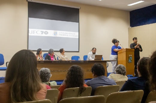 Imagem: A imagem mostra um evento acadêmico em um auditório da Universidade Federal do Ceará (UFC), como indicado pelo logotipo no telão. Há uma mesa com cinco pessoas sentadas, incluindo três mulheres e dois homens. Uma mulher de blusa azul e calça branca está em pé, falando ao microfone em um púlpito à direita, enquanto um intérprete de Libras a acompanha. O público, composto por diversas pessoas, está sentado em cadeiras amarelas, assistindo à apresentação. O ambiente é iluminado por luzes fluorescentes, com paredes em tons neutros e detalhes em azul.