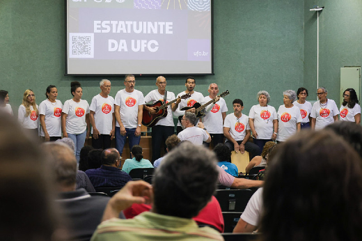 Imagem: A imagem mostra um auditório com um grupo de pessoas vestindo camisetas brancas com um logotipo vermelho no centro. Elas estão de pé na frente da plateia, formando um coral. Dois homens ao centro tocam violão enquanto os demais cantam. O público está sentado e assistindo à apresentação. No fundo, há um telão projetando a frase "ESTATUINTE DA UFC" com um QR code ao lado. O ambiente tem paredes verdes e iluminação artificial.A imagem mostra um auditório com um grupo de pessoas vestindo camisetas brancas com um logotipo vermelho no centro. Elas estão de pé na frente da plateia, formando um coral. Dois homens ao centro tocam violão enquanto os demais cantam. O público está sentado e assistindo à apresentação. No fundo, há um telão projetando a frase "ESTATUINTE DA UFC" com um QR code ao lado. O ambiente tem paredes verdes e iluminação artificial.