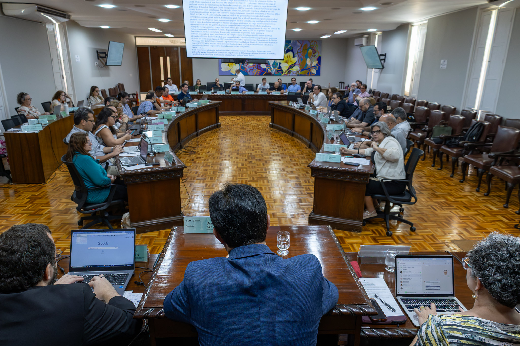 Imagem: A imagem retrata uma reunião formal em uma ampla sala com disposição em semicírculo. Participantes sentados em mesas de madeira usam notebooks e discutem documentos. No centro, um homem de costas usa paletó azul. Um telão projeta um texto, e ao fundo há um painel colorido. O ambiente tem piso de madeira e poltronas dispostas ao redor. (Foto: Guilherme Silva/UFC Informa)