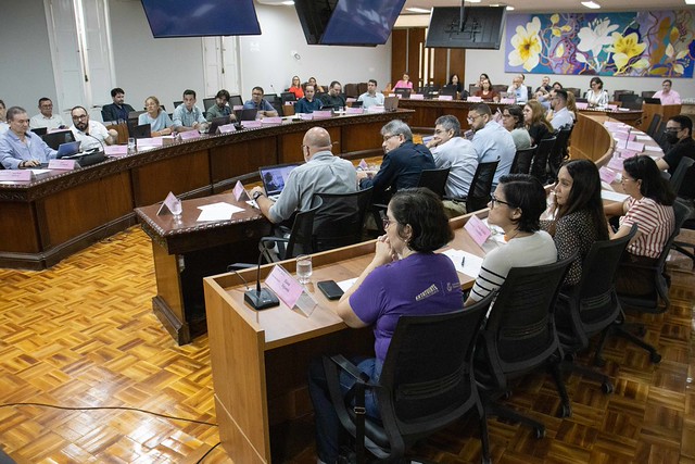 Imagem: Homem de costas, vestindo terno azul, observa demais integrantes do conselhos. Todos estão sentados em uma bancada em formato circular (Foto: Viktor Braga/UFC)