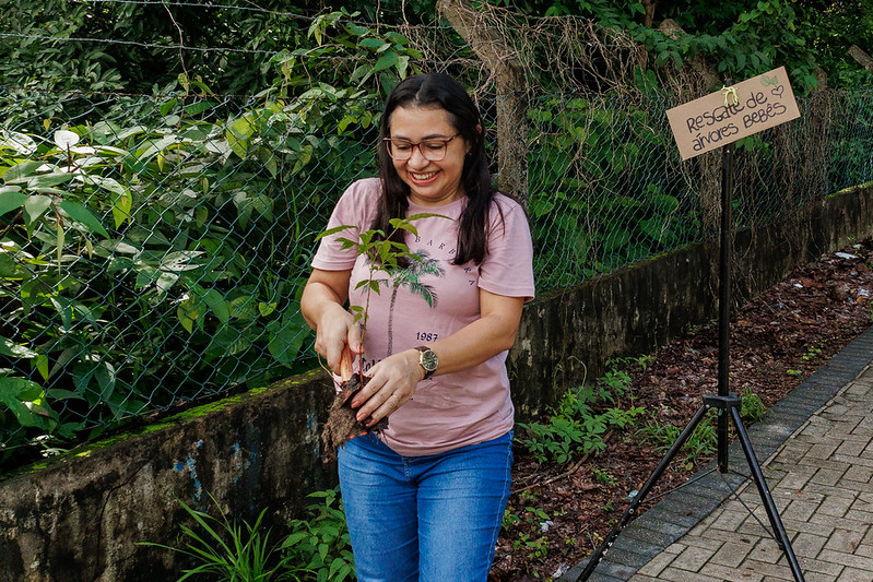 Imagem: estudante mulher, usando camiseta rosa e óculos, segura muda de árvore gonçalo-alves com pá, no entorno da Matinha do Pici, no Campus do Pici da UFC.