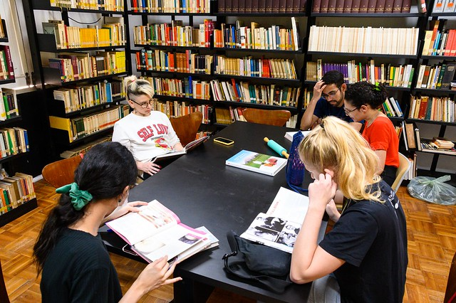 Imagem: Sala de biblioteca com estantes altas repletas de livros organizados ao fundo e nas laterais. No centro, uma mesa retangular preta reúne cinco estudantes sentados, concentrados na leitura e consulta de materiais. Uma estudante de cabelos escuros presos por um laço verde folheia um livro aberto. À frente, outra estudante loira consulta um livro, apoiando uma das mãos no rosto. Ao lado, dois estudantes observam o mesmo material, enquanto uma estudante de cabelos claros, sentada ao fundo, lê um livro com atenção. Sobre a mesa, há livros, cadernos, estojo e garrafas. O ambiente é iluminado, silencioso e organizado, sugerindo momento de estudo coletivo e pesquisa acadêmica