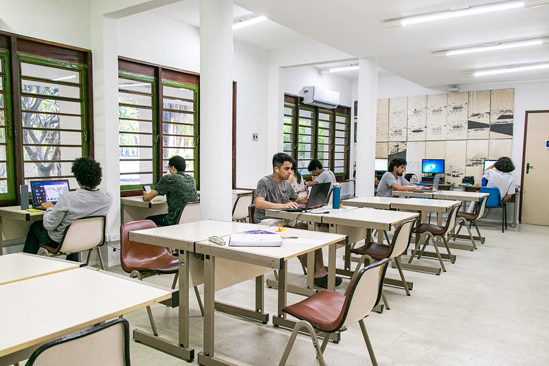 Imagem: Sala de biblioteca com três janelas e 20 mesas pequenas, com cadeiras, e sete alunos, cada um em uma mesa, estudando em computador e celular. A sala tem teto branco, três colunas partindo do teto, ar condicionado e porta no lado direito.