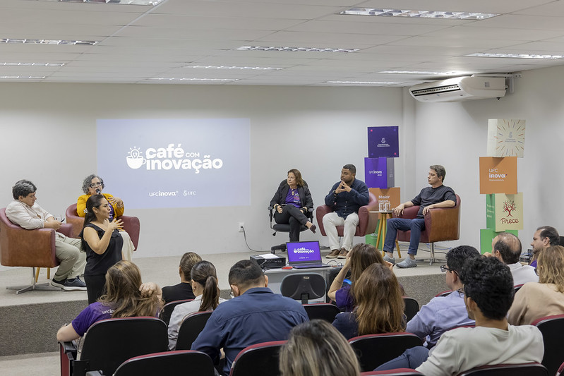 Imagem: Roda de conversa com quatro palestrantes e uma mediadora, sentados em cadeiras de cor vermelha e com pés de madeira. Projetada de maneira centralizada na parede do auditório, há o logotipo do evento, intitulado “Café com Inovação”, na cor branca. Há, ainda, na cena uma intérprete de Libras usando roupa na cor preta. Parte da plateia do evento também é mostrada na parte inferior direita da imagem, sentada nas cadeiras do auditório.