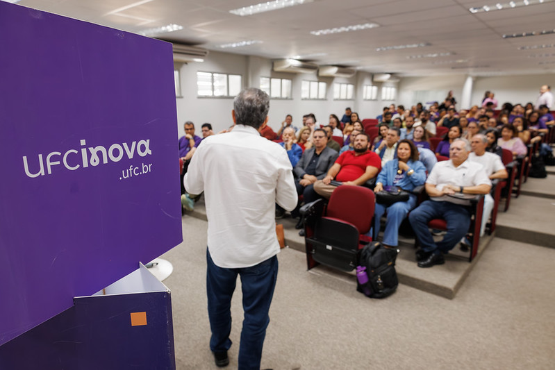 Imagem: Auditório com plateia cheia durante palestra de um ano da Agência UFC Inova, com um cubo na cor roxa em destaque no lado esquerdo da foto, com a inscrição “ufcinova.ufc.br”.