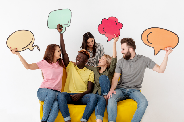 Trata-se de uma foto de cinco pessoas sentadas em um sofá amarelo, sorrindo e segurando balões coloridos em formato de diálogo. Ao fundo, uma parede branca, sugerindo comunicação, troca de ideias e trabalho em equipe.