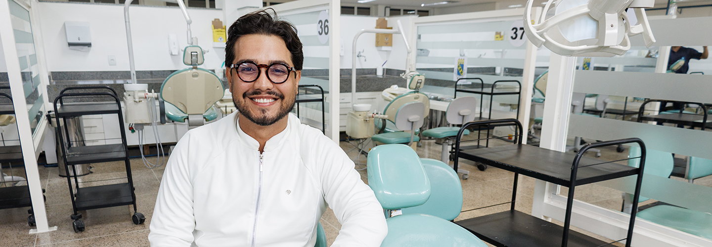 Estudante do curso de Odontologia posa em clínica odontológica tendo ao fundo equipamentos da área